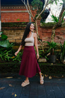 Woman in a Wildalaya Everyday Hemp Wrap Skirt side view, relaxed fit, eco-friendly hemp textile mulberry skirt and beige top standing in a garden with plants and a brick wall in the background.