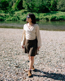 A woman wearing a Wildalaya beige wrap-style long-sleeve top and a dark brown skirt walks on a rocky riverbank with trees and water in the background.
