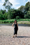 A woman wearing a black sleeveless midi dress and sunglasses stands on a gravel riverbank with trees and water in the background.