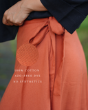 Close-up of a person wearing a Wildalaya Red clay cotton wrap skirt with a tie waist, paired with a dark navy top. A circular fabric texture swatch is overlaid on the left thigh area with white text reading '100% COTTON AZO-FREE DYE NO SYNTHETICS'.