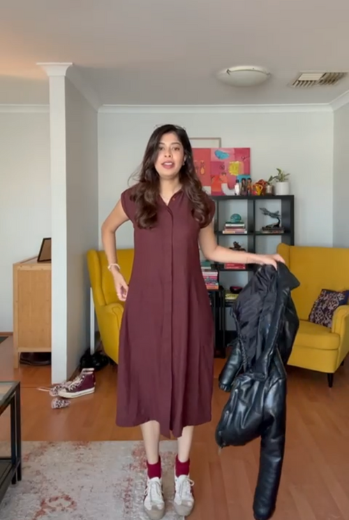 Woman in a hemp maroon dress holding a black jacket in a room with yellow chairs and a bookshelf.
