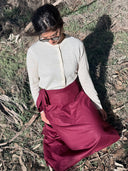 A woman sitting outdoors on dry grass and branches, wearing a cream long-sleeve button-front top and a deep mULBERRY HEMP wrap skirt.