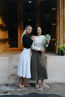 Two women standing on a steps in front of a wooden building, one holding flowers wearing Wildalaya Wrap Skirts in moonstone white and mossy grove green 